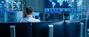 image of the back of a professional man sitting in a blue lit room of computers and screens with maps and graphs
