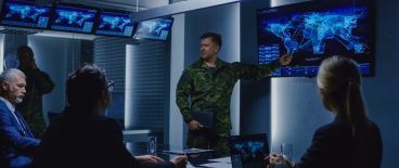 Image of military manpointing at computer screen as he briefs professionals sitting at tables