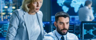 Image of woman standing and man sitting in front of a computer screen in a blue lit room or network operating center