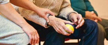 image of elderly man sitting with small rubber ball in his hand, with a helper at his side