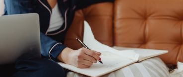 person sitting on couch with open laptop on lap and writing in notebook