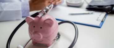 A stethoscope placed on top of a piggy bank with a doctor in the background using a phone